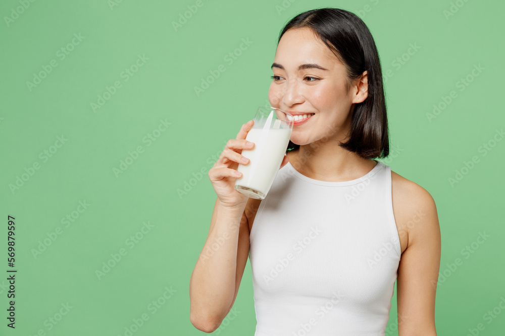 Young smiling happy fun woman wear white clothes hold in hand glass drink milk look aside isolated on plain pastel light green background. Proper nutrition healthy fast food unhealthy choice concept. Young smiling happy fun woman wear white clothes hold in hand glass drink milk look aside isolated on plain pastel light green background. Proper nutrition healthy fast food unhealthy choice concept.