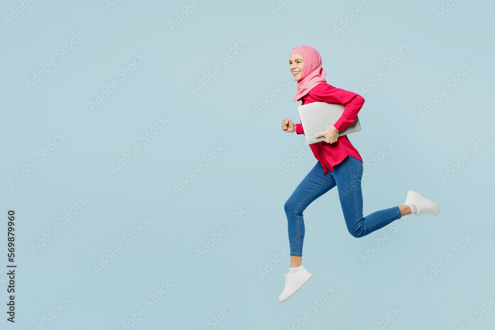 Full body side profile view young arabian asian muslim IT woman wear pink abaya hijab jump high hold laptop pc computer isolated on plain blue cyan background studio portrait People uae islam concept. Full body side profile view young arabian asian muslim IT woman wear pink abaya hijab jump high hold laptop pc computer isolated on plain blue cyan background studio portrait People uae islam concept.