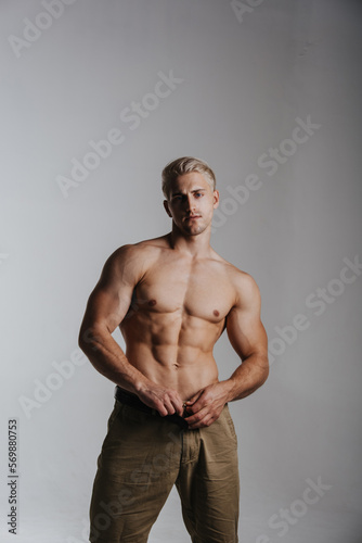 Handsome male model wearing sand pants. Sexy shirtless man in studio. Blond muscular guy standing on light grey background.