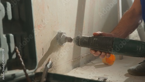 Man drilling round hole in concrete wall for socket. Builder drills hole with rotating electric drill or perforator. White dust from plaster crumbles. Hands and power tool close up.