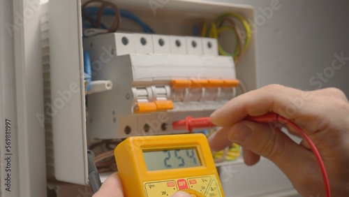 Work under voltage, electrician is checking serviceability of equipment, measuring voltage by yellow multimeter. Close up of a mans hands testing white switch box check voltage switchboard.