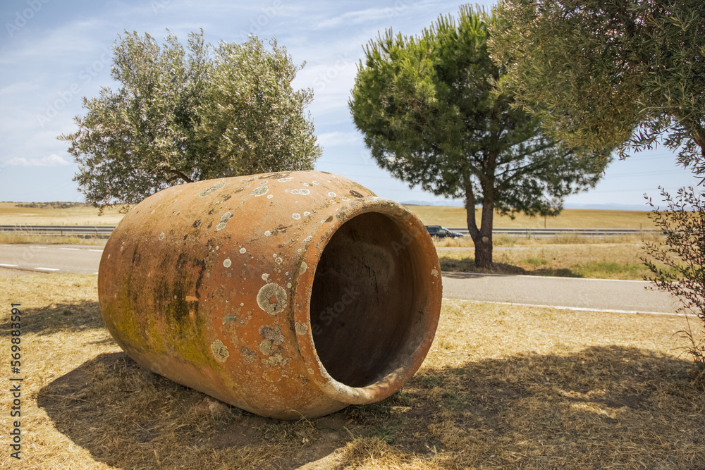 A large ceramic vessel for storing olive oil and other products is located on the ground against the background of olive trees in the field. Mediterranean culture. Spain. A large ceramic vessel for storing olive oil and other products is located on the ground against the background of olive trees in the field. Mediterranean culture. Spain.