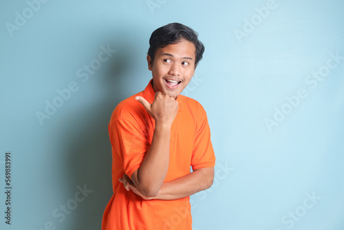 Portrait of attractive Asian man in orange shirt with thumb pointing away on empty space. Advertising concept. Isolated image on blue background