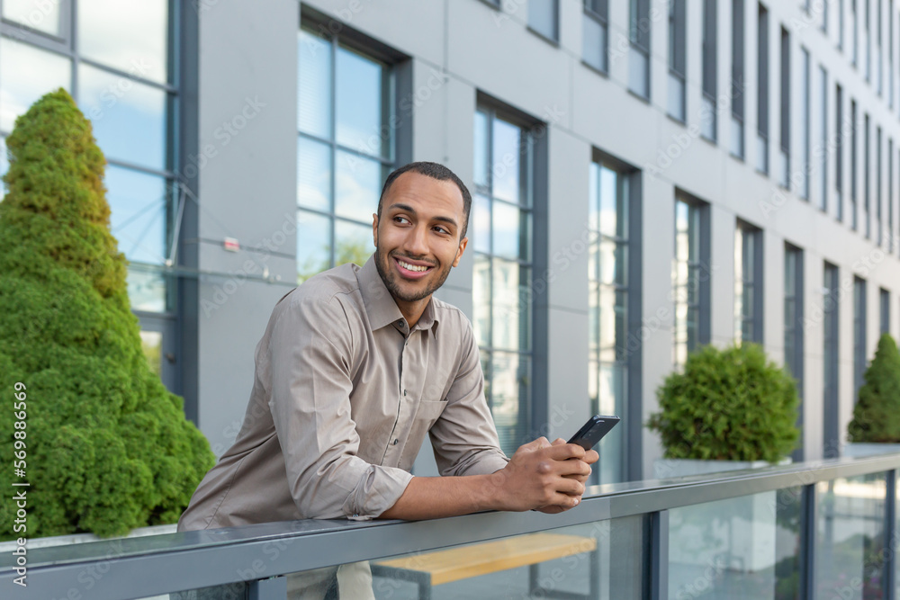 Cheerful young student in shirt using phone outside university campus, african american man smiling and looking away, holding smartphone, typing message and making call.