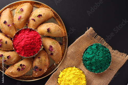 Traditional indian sweet mithai mawa gujia pirukiya with dry powder color gulal abeer during Holi Indian festival of colors presented on a brass plate bowl isolated on black background photo