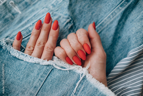 Beautiful female hands with a bright red matte manicure on a background of denim. Stylish oval nail design. Summer manicure. Copy space.