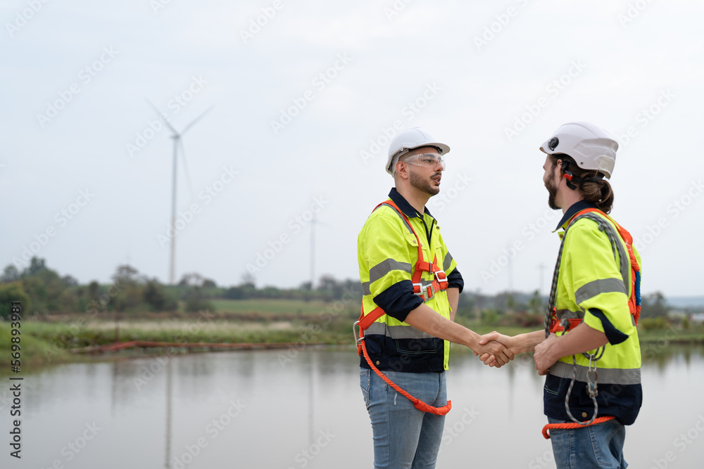 Two male engineers windmill in uniform and helmet safety shaking hands after to work success in wind turbine farm to generate electrical energy, Renewable power energy. Two male engineers windmill in uniform and helmet safety shaking hands after to work success in wind turbine farm to generate electrical energy, Renewable power energy.
