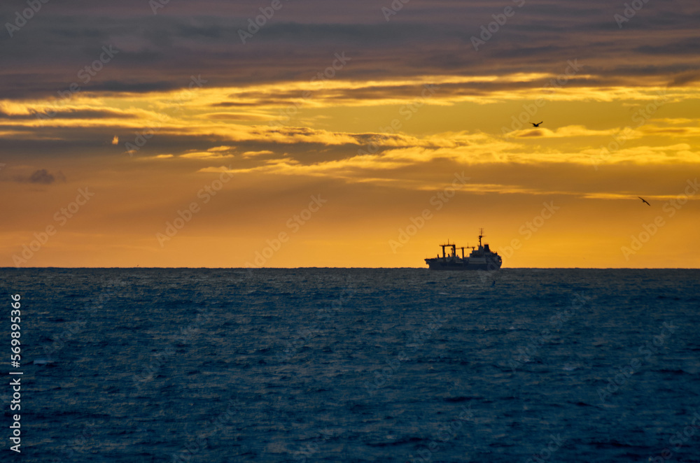 Silhouette of a fishing ship on the horizon of the sea against the backdrop of a dramatic sunset with sunbeams