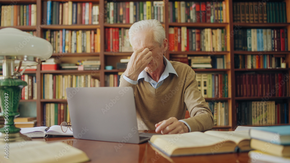 Senior man translator rubbing tired eyes, working long hours on laptop, writer Senior man translator rubbing tired eyes, working long hours on laptop, writer