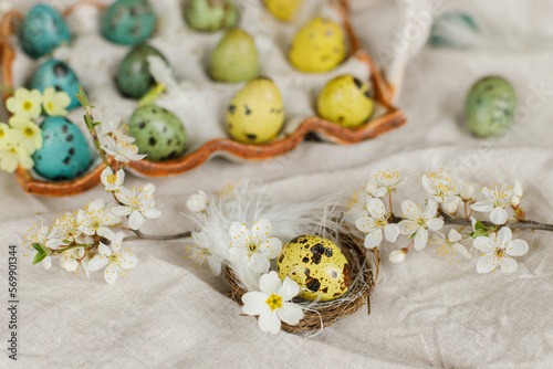 Happy Easter! Stylish easter eggs and blooming spring flowers on rustic table. Natural painted quail eggs in tray, feathers and cherry blossoms on linen fabric. Rustic easter still life