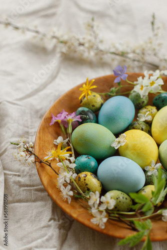 Rustic easter still life. Stylish easter eggs and blooming spring flowers in wooden bowl on linen fabric. Happy Easter! Natural painted eggs and blossoms on rural table