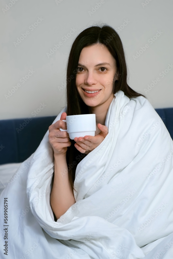 Attractive brunette woman wrapped in a white blanket smiling with cup of tea or coffee in the morning, vertical video. She is sitting on bed and looking at camera. Lazy morning, good mood concept. Attractive brunette woman wrapped in a white blanket smiling with cup of tea or coffee in the morning, vertical video. She is sitting on bed and looking at camera. Lazy morning, good mood concept.