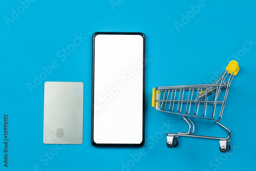 Card, mobile phone with white screen, shopping cart on blue background, copy space, top view. Online shopping and payment concept