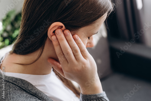Close up of young brunette female holding painful ear  suddenly feeling strong ache. Unhealthy caucasian woman 20s suffering from painful otitis sitting on couch at home. Health problems concept