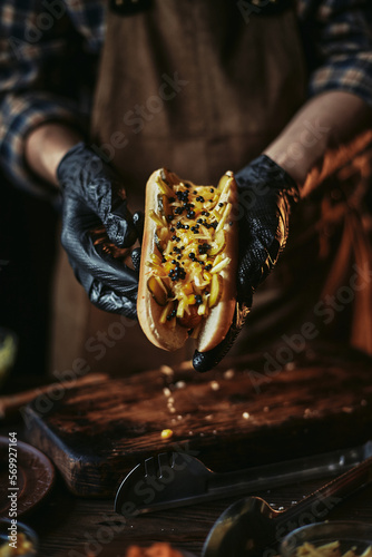 The chef in gloves holds a hot dog. Fast food cooking. Classic hot dog in dark style.