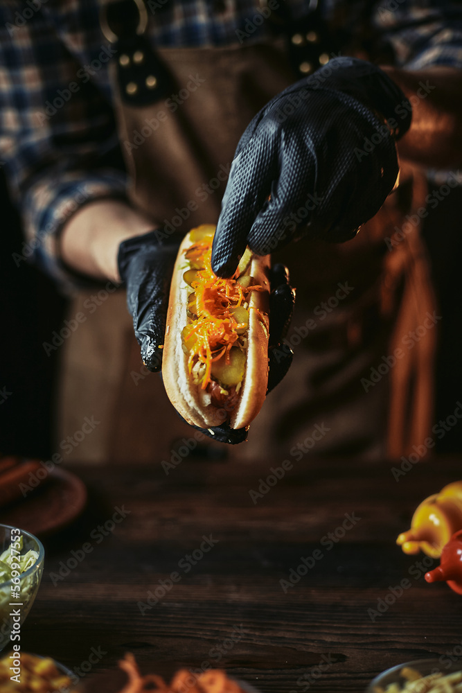 A guy in a leather apron makes a hot dog. The chef in black gloves puts a carrot in a bun. A guy in a leather apron makes a hot dog. The chef in black gloves puts a carrot in a bun.