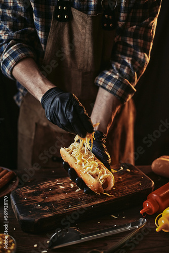 A guy in an apron and gloves sprinkles cheese on a hot dog. Hot dog cooking. Creating classic fast food.