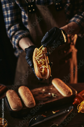 A guy in a leather apron makes a hot dog. The chef in black gloves puts french fries in a bun.