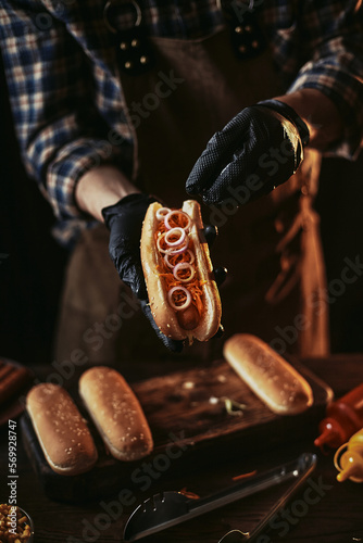 A guy in a leather apron makes a hot dog. A chef in black gloves puts an onion in a bun.