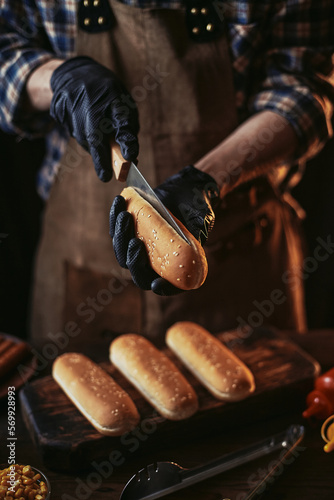 A guy in a leather apron cuts a hot dog bun. Photo of the process of creating a hot dog. Cooking food in the kitchen.