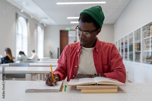 Valokuva Talented young black guy scientist wearing glasses studying in library, reading book and taking notes