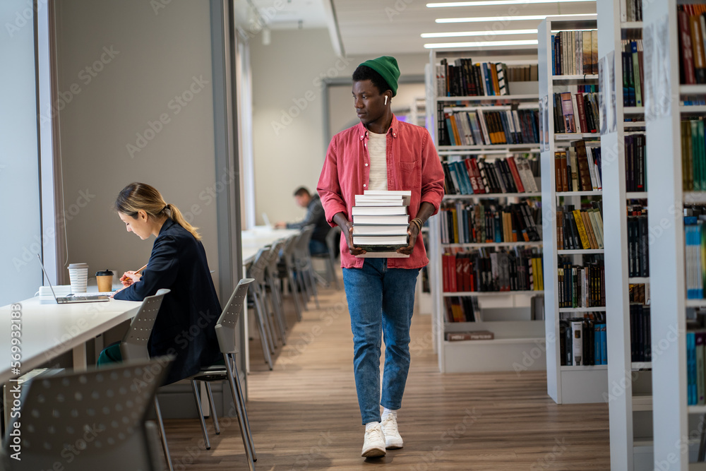 Bookworm. Full length of African American international student guy walking with stacked books through college library. Male librarian organizing and managing resources. Education concept