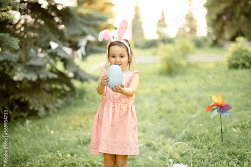 Little cute girl wear bunny ears holding big blue painted egg on Easter egg hunt in park