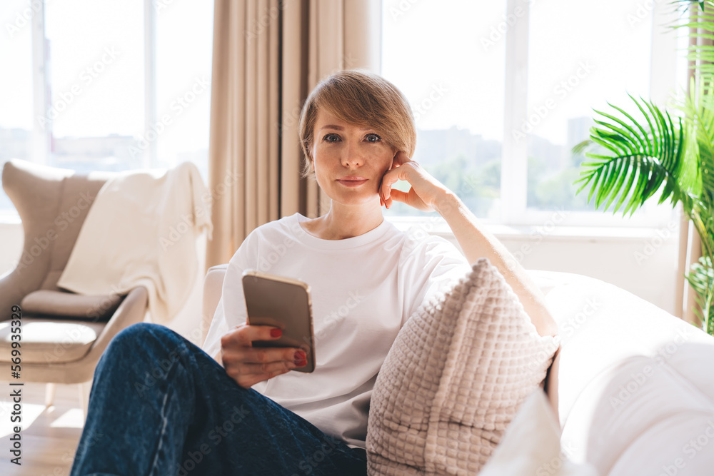 Pensive mature woman sitting on sofa with smartphone and looking at camera Pensive mature woman sitting on sofa with smartphone and looking at camera