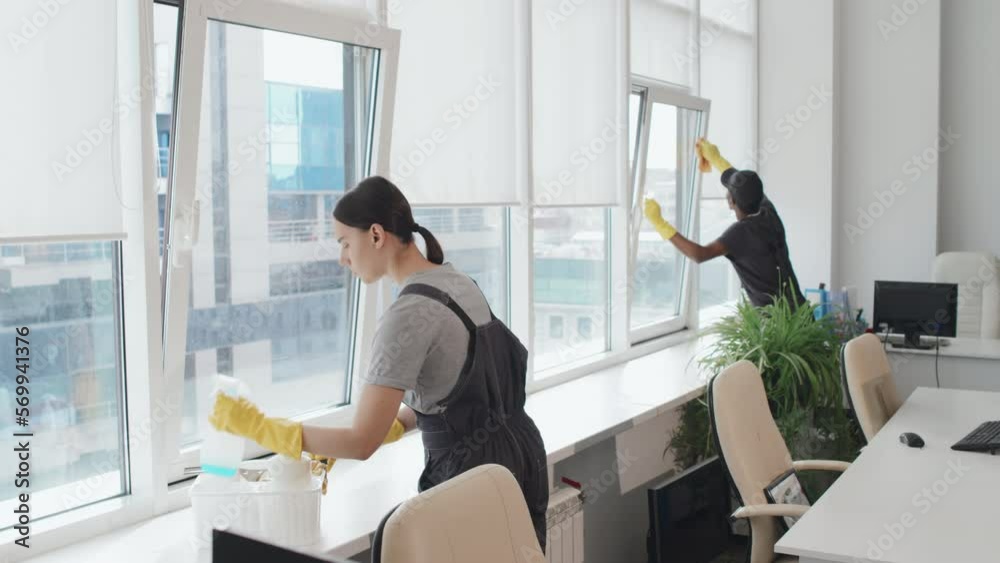 Slow motion of young Black man and Caucasian woman wearing uniform with yellow rubber gloves cleaning windows in modern office using detergent and tools