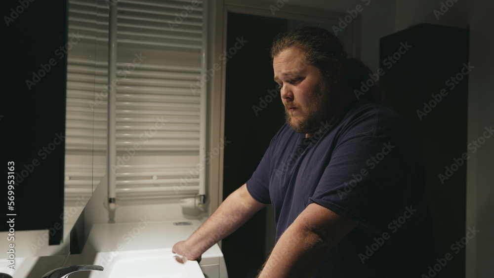 Sad young man standing in bathroom concerned about problems with pensive expression. A male overweight person in front of mirror