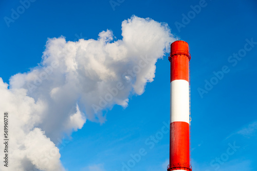 Red-white pipe of a plant with exhaust white smoke. Blue sky.