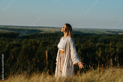 Happy Young Woman Meditating and Catching Sunlight Outdoors at Sunset with Scenic Landscape on the Background