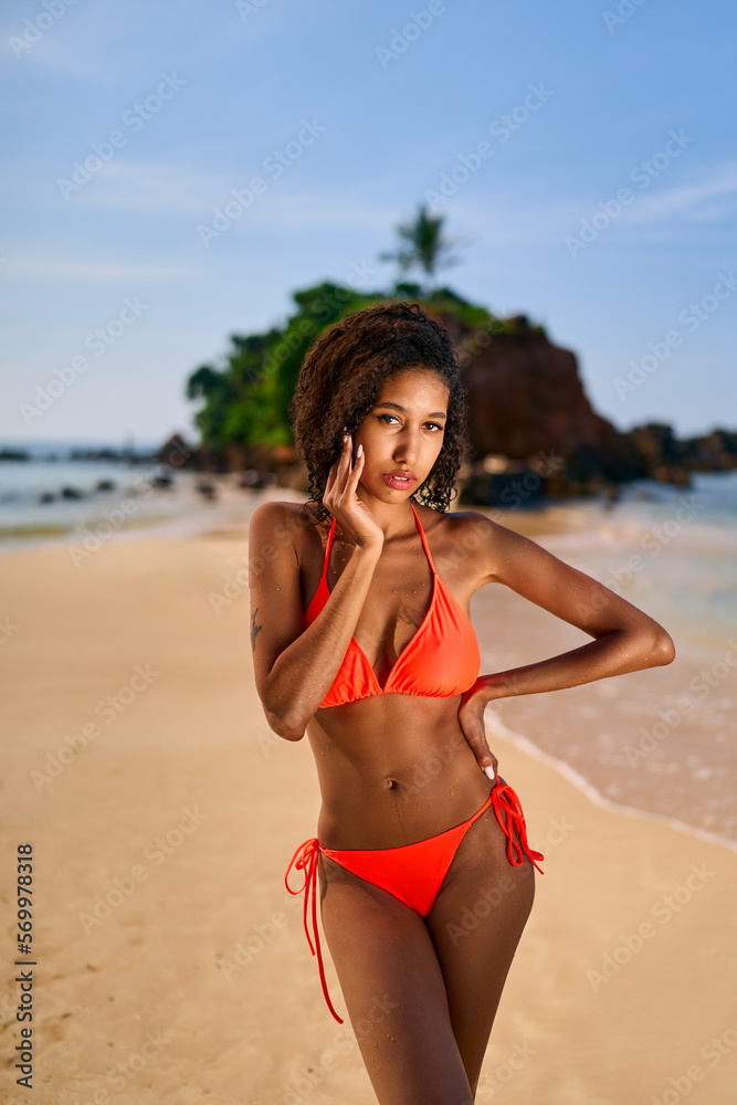 Portrait of serene looking fit attractive young black female posing in bikini at exotic beach by ocean. African model posing against tropical island background by sea. Fashion portrait of black woman