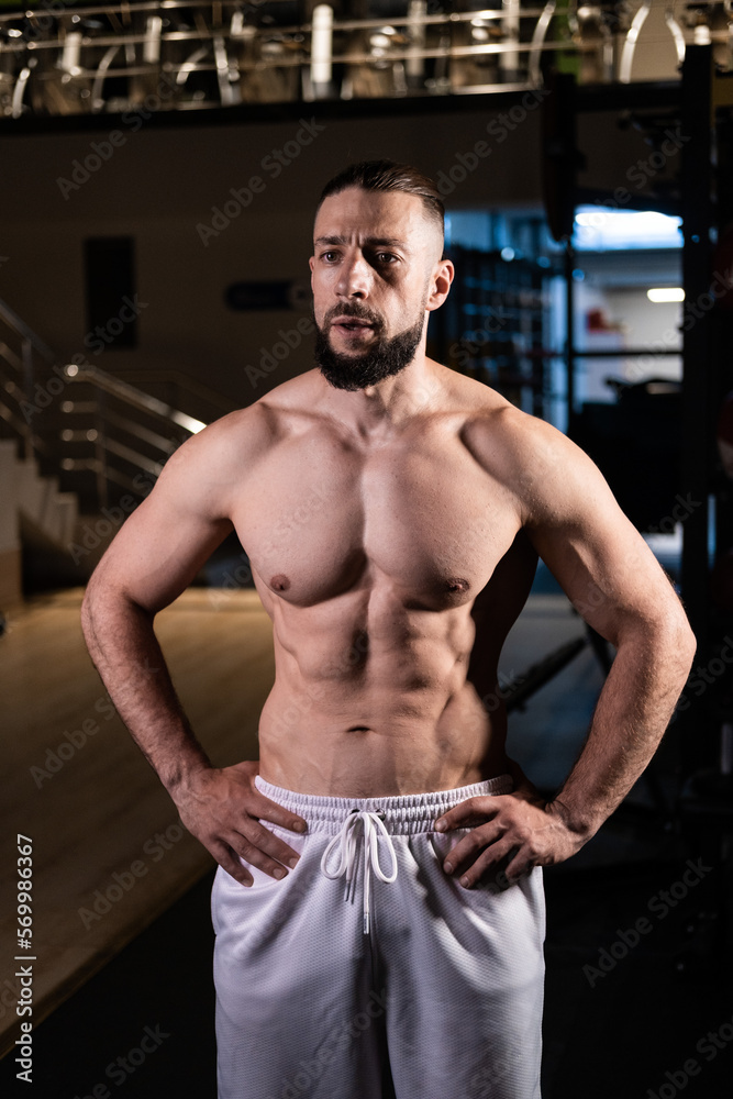 Fototapeta premium A handsome muscular man with a spectacular body in white pants in the gym. Sports, healthy lifestyle