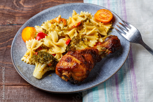 Pasta with vegetables and cherry tomatoes next to a baked chicken leg on the table next to a light napkin and a fork.