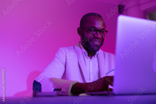 a black man in a white suit and glasses works at a laptop. a man in glasses works in the office in the evening