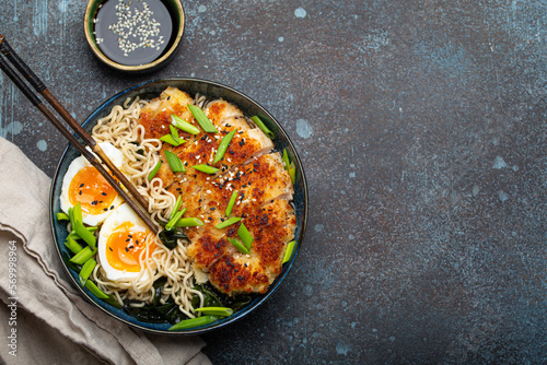 Asian noodles ramen soup with deep fried panko chicken fillet and boiled eggs in ceramic bowl with chop sticks and soy sauce on stone rustic background top view, space for text