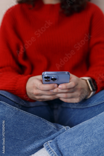 Young girl typing message on smart phone. Close up photo of unrecognizable female person sitting on couch and using mobile phone