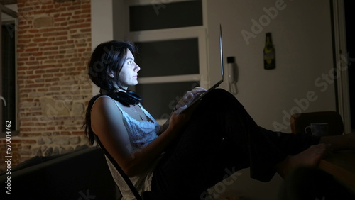 Woman working at night at home in front of laptop computer in the dark. Computer screen glowing on person face while browsing internet online