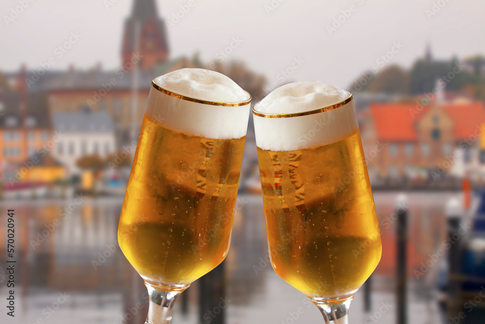 Pair of beer glasses making a toast with old German city blurred in background. Cold refreshing ale with white foam. Pair of beer glasses making a toast with old German city blurred in background. Cold refreshing ale with white foam.
