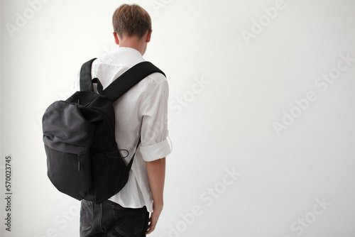 The guy with a bagpack on his shoulders is poaing  on a white background. The teenager  is wearing a white T-shirt and black jeans photo