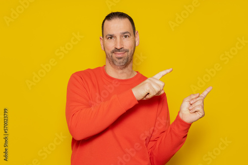 Smiling happy man 40s in casual red pullover pointing index fingers to the side in mock-up workspace area isolated on yellow colored background studio portrait. People lifestyle concept