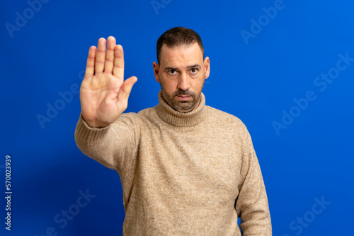 Hispanic handsome man with beard wearing a beige turtleneck sweater over isolated blue background doing stop gesture.