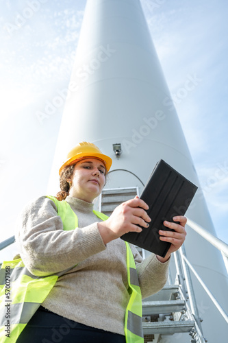 Female engineer with brown curly hair and yellow helmet using her digital tablet while standing in front of a wind turbine during daylight, low angle view