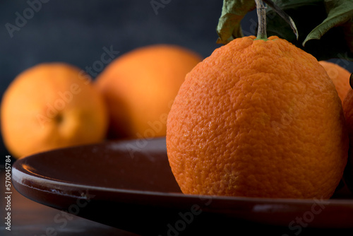 Detalle de una naranja en un plato de ceràmica marrón. Al fondo, fuera de foco, otras naranjas. photo