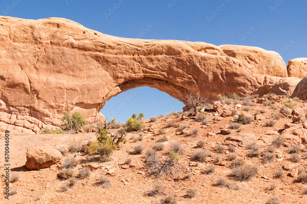 Natural sandstone arch formation and sagebrush in the desert of Arches National Park Utah with a cloudless blue sky.