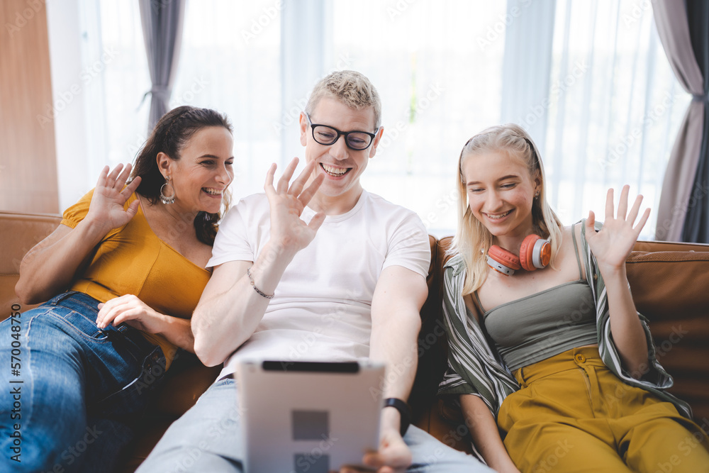 Smiling young family of father with spectacles sitting on couch with wife and daughter wearing headphones around neck at home waving hand on digital tablet screen during online video call