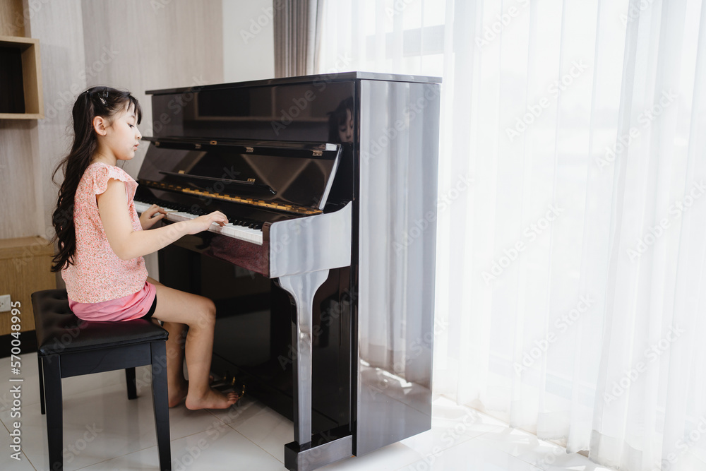 Cute and focused young little Asian girl practicing songs and melody while pressing white and black key and creating new music on piano as hobby and dedication at home Cute and focused young little Asian girl practicing songs and melody while pressing white and black key and creating new music on piano as hobby and dedication at home