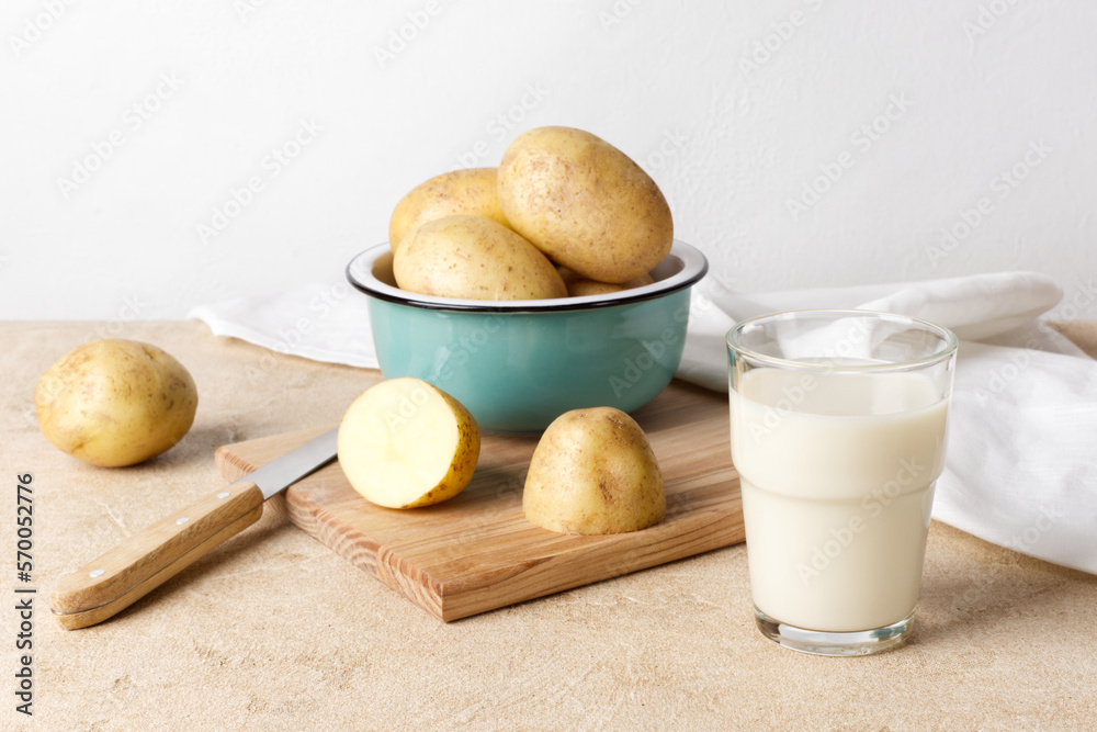 Potato milk, poured into a glass, stands next to a ceramic bowl containing several unpeeled potatoes. Close-up, horizontal orientation. Potato milk, poured into a glass, stands next to a ceramic bowl containing several unpeeled potatoes. Close-up, horizontal orientation.