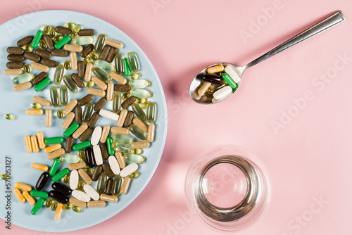 Many dietary supplements tablets, glass of water and spoon for daily pills intake on pink background. Set of zinc pills, antioxidants from aging, lecithin and adaptogen.